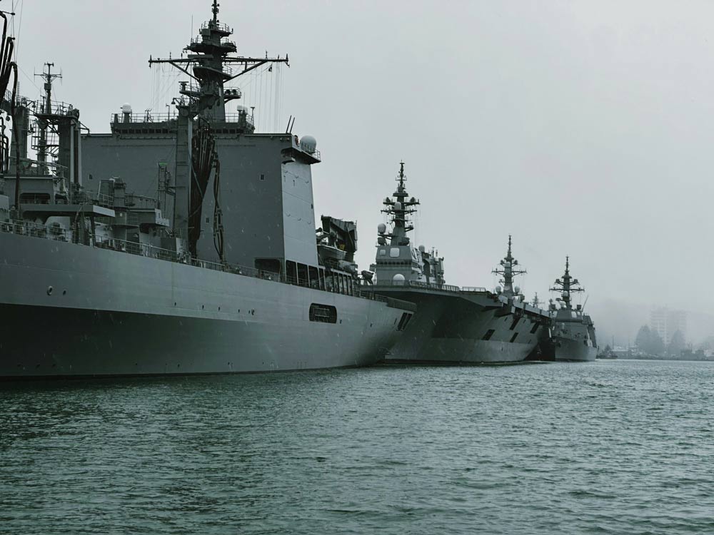 Three naval ships docked side by side on a body of water with a cloudy sky.