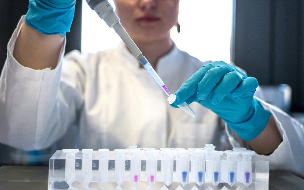Person in a lab setting with gloves on, holding a pipette over test tubes.